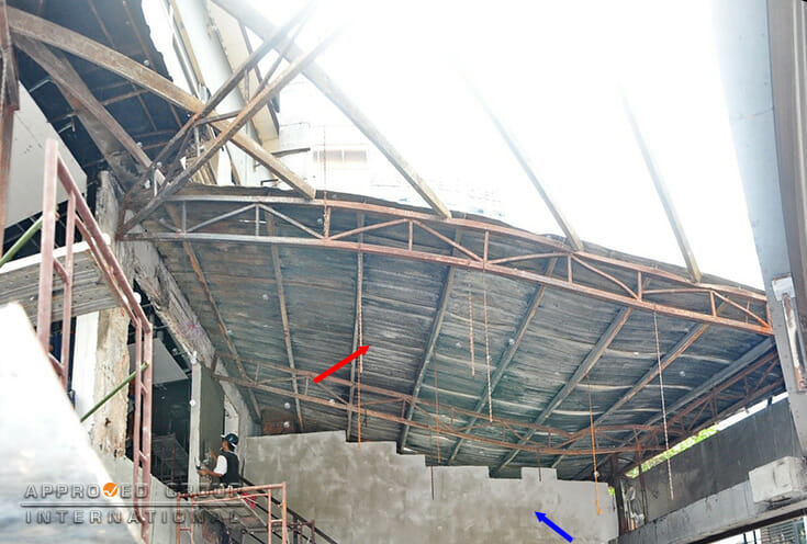 Figure 1: General view of the extent of fire damage onto the roof of the Restaurant B (red arrow) and the newly erected cement wall on top of the pre-existing low-level wall after the fire (blue arrow).